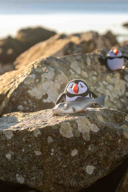 Plush Puffin soft toy with a fish on a rocky surface beside the shore.