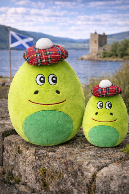 Two green plush nessie soft toys with tartan hats on a stone wall with a scenic background.