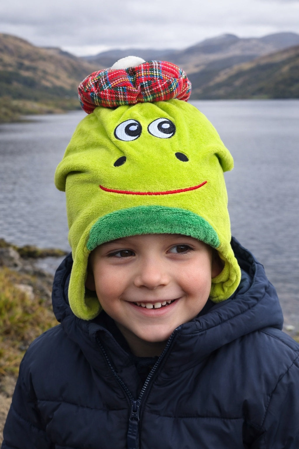 Child wearing a green nessie hat with a tartan pattern on top, standing in front of a lake with mountains in the background.