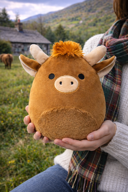 Person holding a brown plush Highland cow soft toy outdoors with mountains in the background.