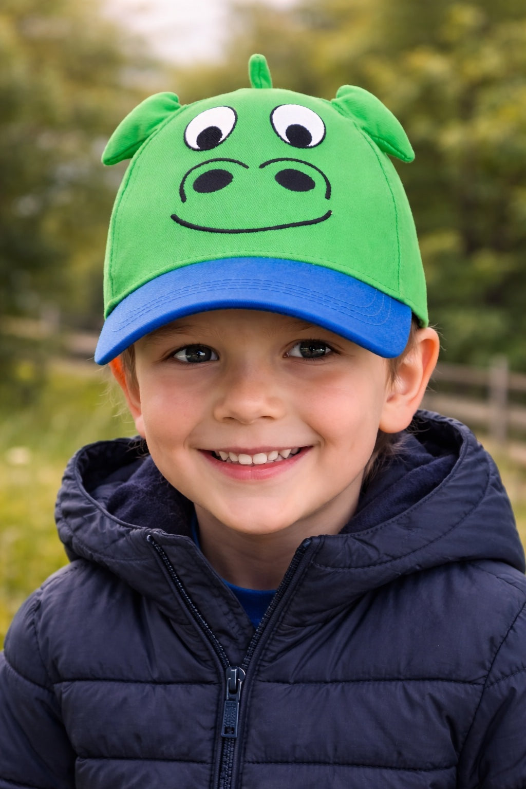 Child wearing a green and blue nessie cap with a cartoon face design outdoors.