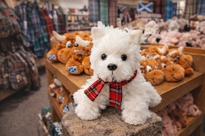Westie dog soft toy with a red tartan scarf on a stone block in a store setting.